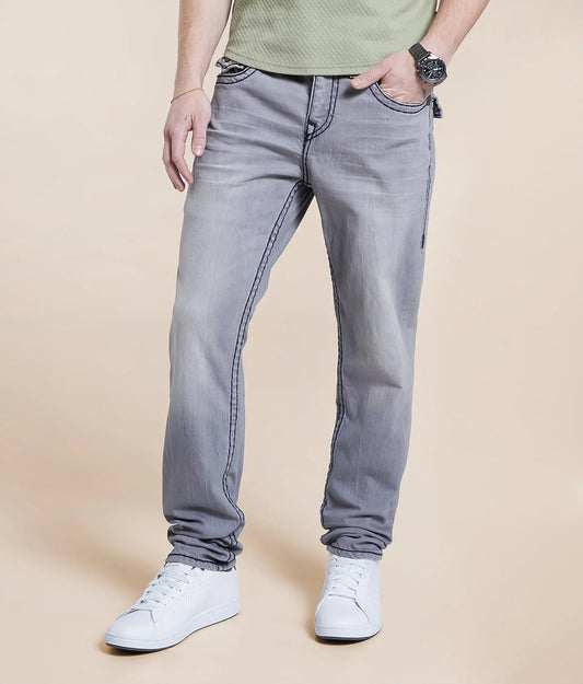 Someone wearing dirtylabels Grey Relaxed Skinny Stretch Jeans, a green shirt, and white sneakers stands against a beige background. Their left hand is in the pocket and they sport a black watch. The photo is cropped at the shoulders.