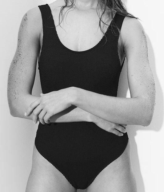 A person wearing the dirtylabels Padded-cup High-leg swimsuit in textured black stands confidently with crossed arms and wet hair against a neutral backdrop. The high-cut design and texture are accentuated in this black-and-white photo, emphasizing their assured stance.